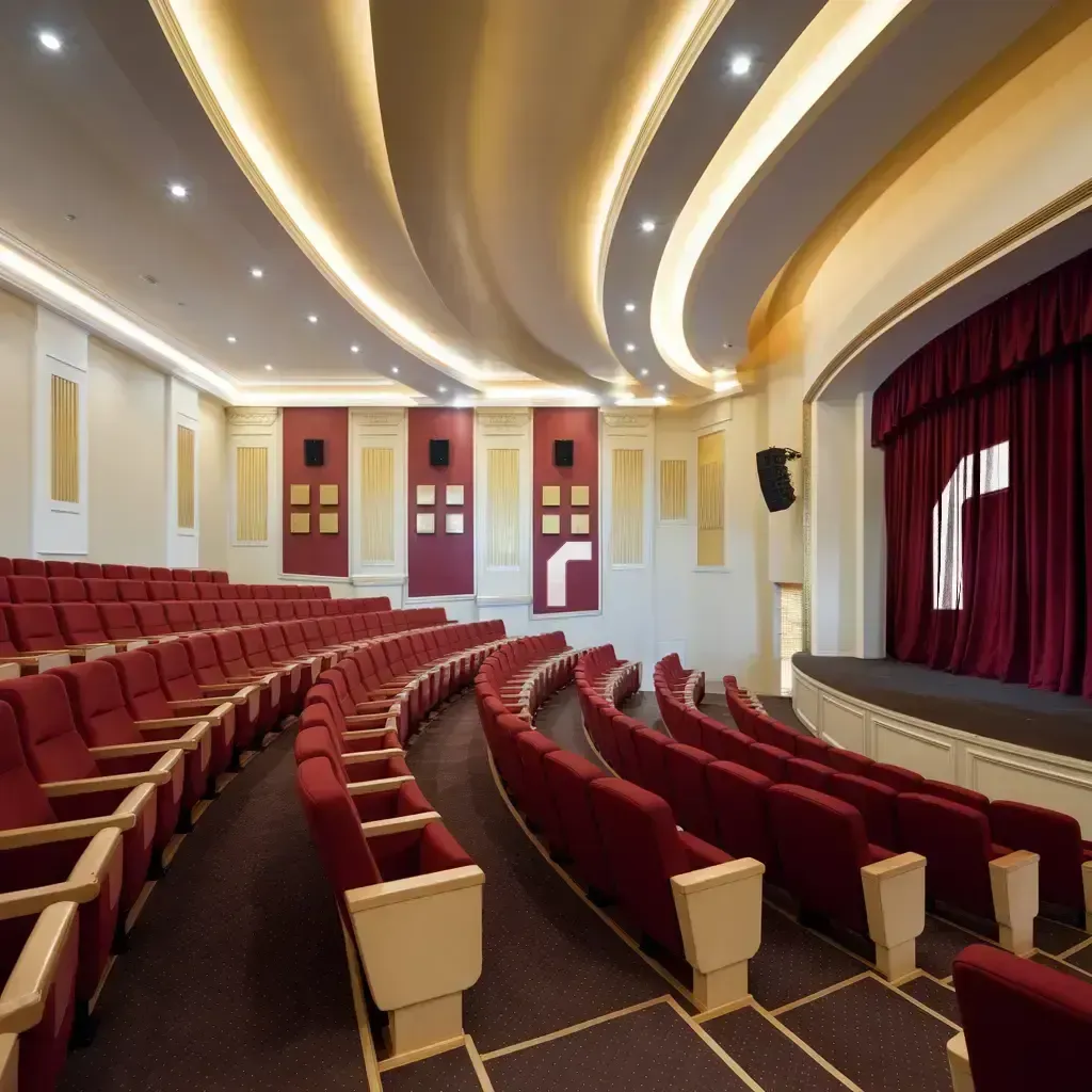 Ruqaqa-fitted theater hall with tiered red seating, a crimson stage curtain, acoustic wall panels, mounted speakers and curved cove ceiling lighting